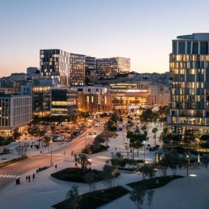 Vue au crépuscule d'un quartier moderne de Marseille, avec des immeubles vitrés illuminés et une place animée, reflétant l'atmosphère vivante et cosmopolite de la ville.