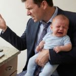 Entrepreneur en télétravail habillé en costume et pyjama, l'air stressé avec un bébé qui pleure dans les bras, illustrant le manque de frontières entre vie de famille et travail à domicile.