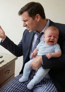 Entrepreneur en télétravail habillé en costume et pyjama, l'air stressé avec un bébé qui pleure dans les bras, illustrant le manque de frontières entre vie de famille et travail à domicile.